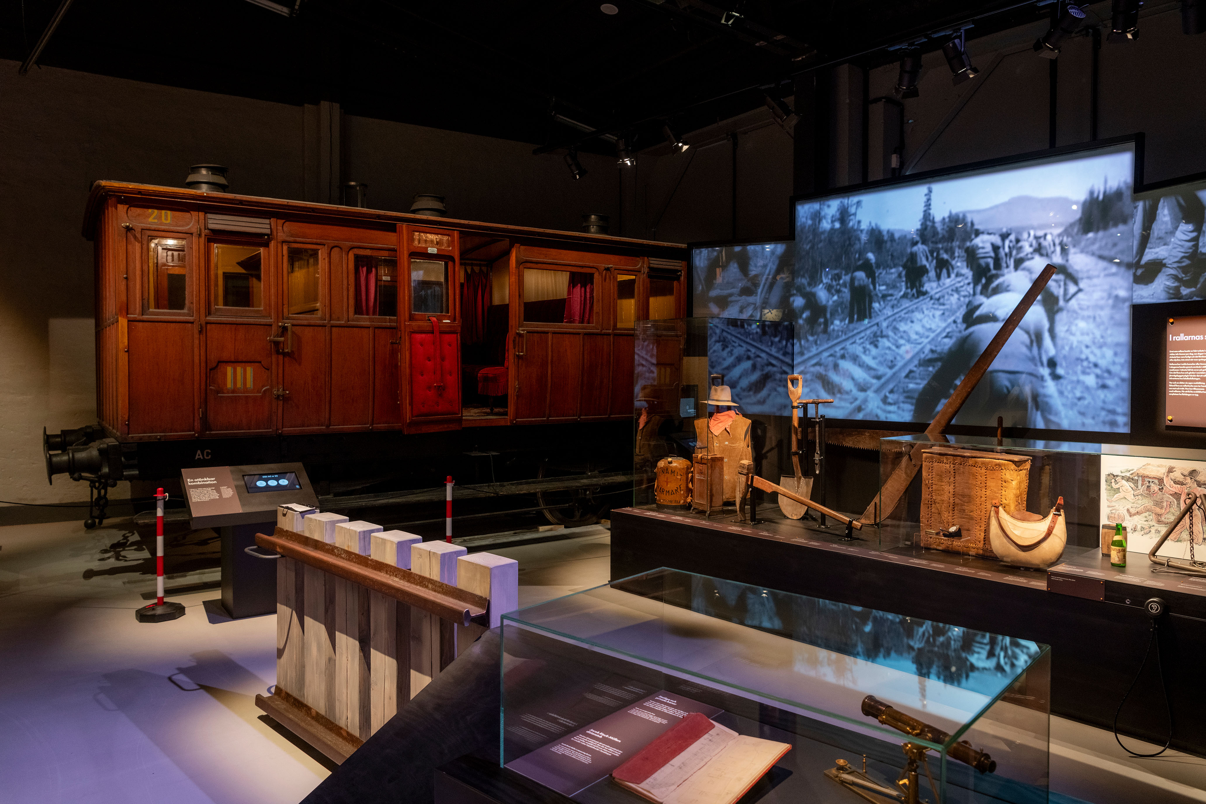 A historical carriage, a exhibition stand and a screen with a black and white film showing is standing in an exhibition hall.