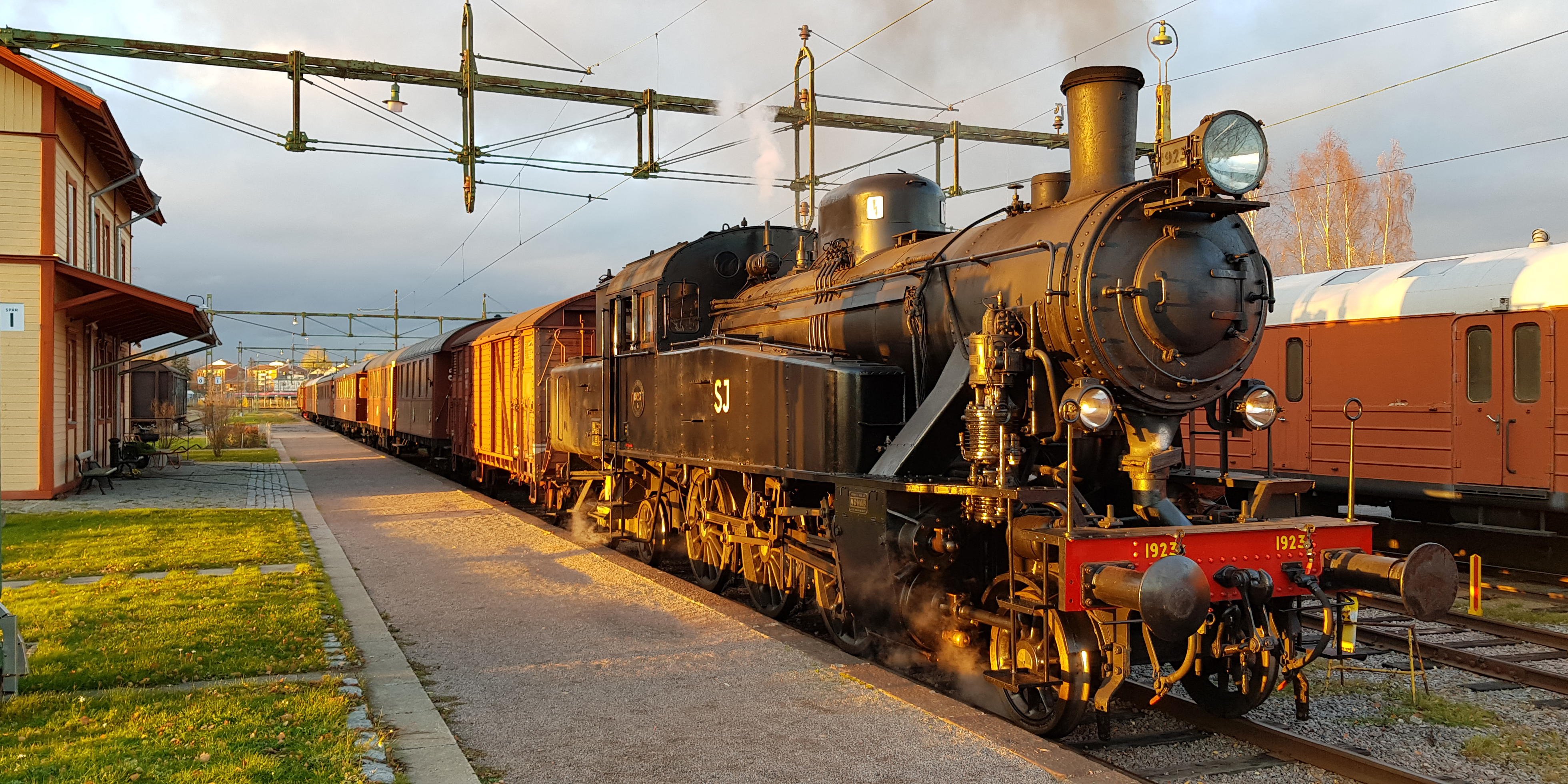 Locomotive standing at the platform in front of the station building, puffing smoke in dusk.