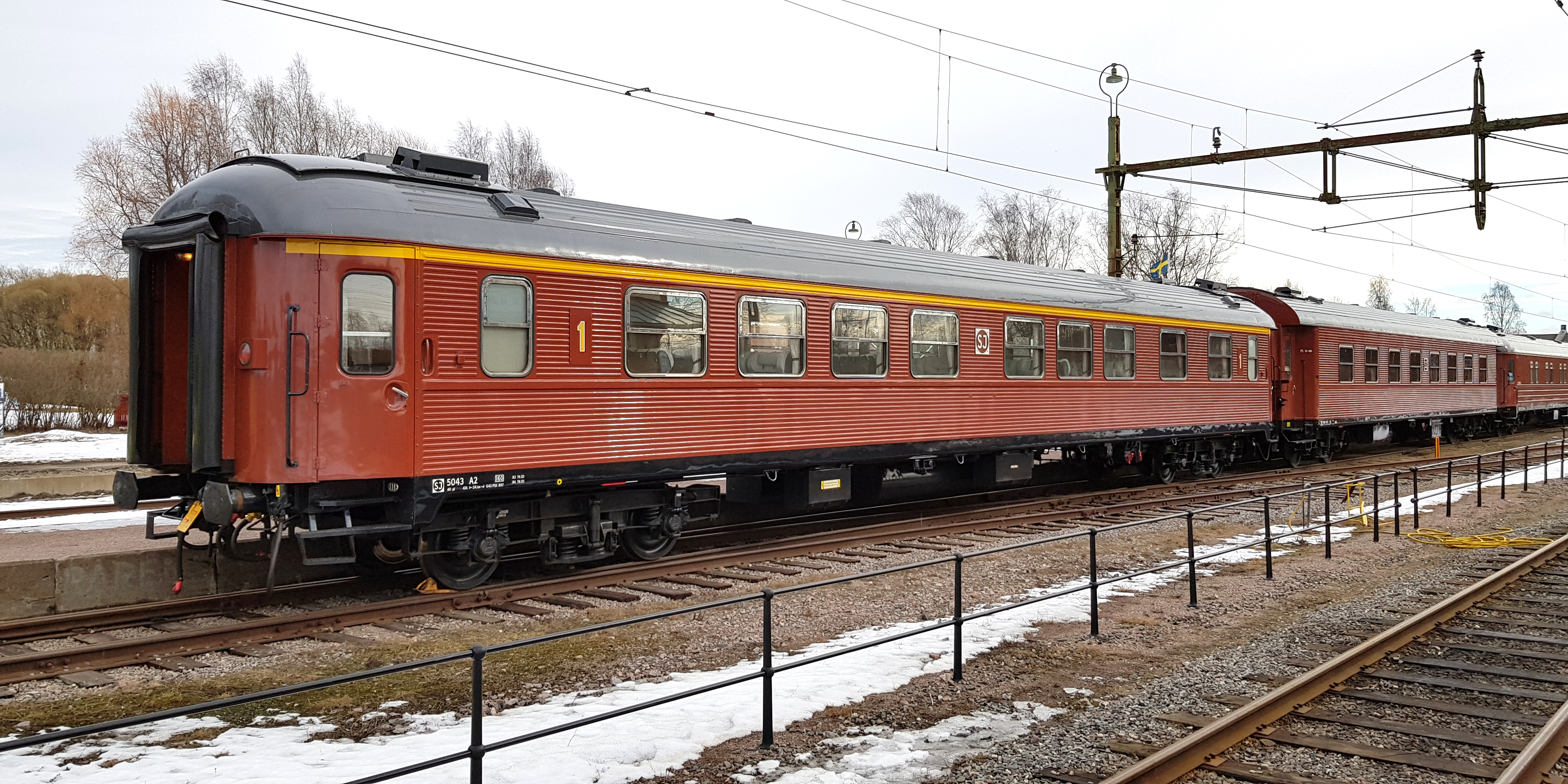 Rust red passenger car with yellow stripe above the windows connected last in line of carriages.