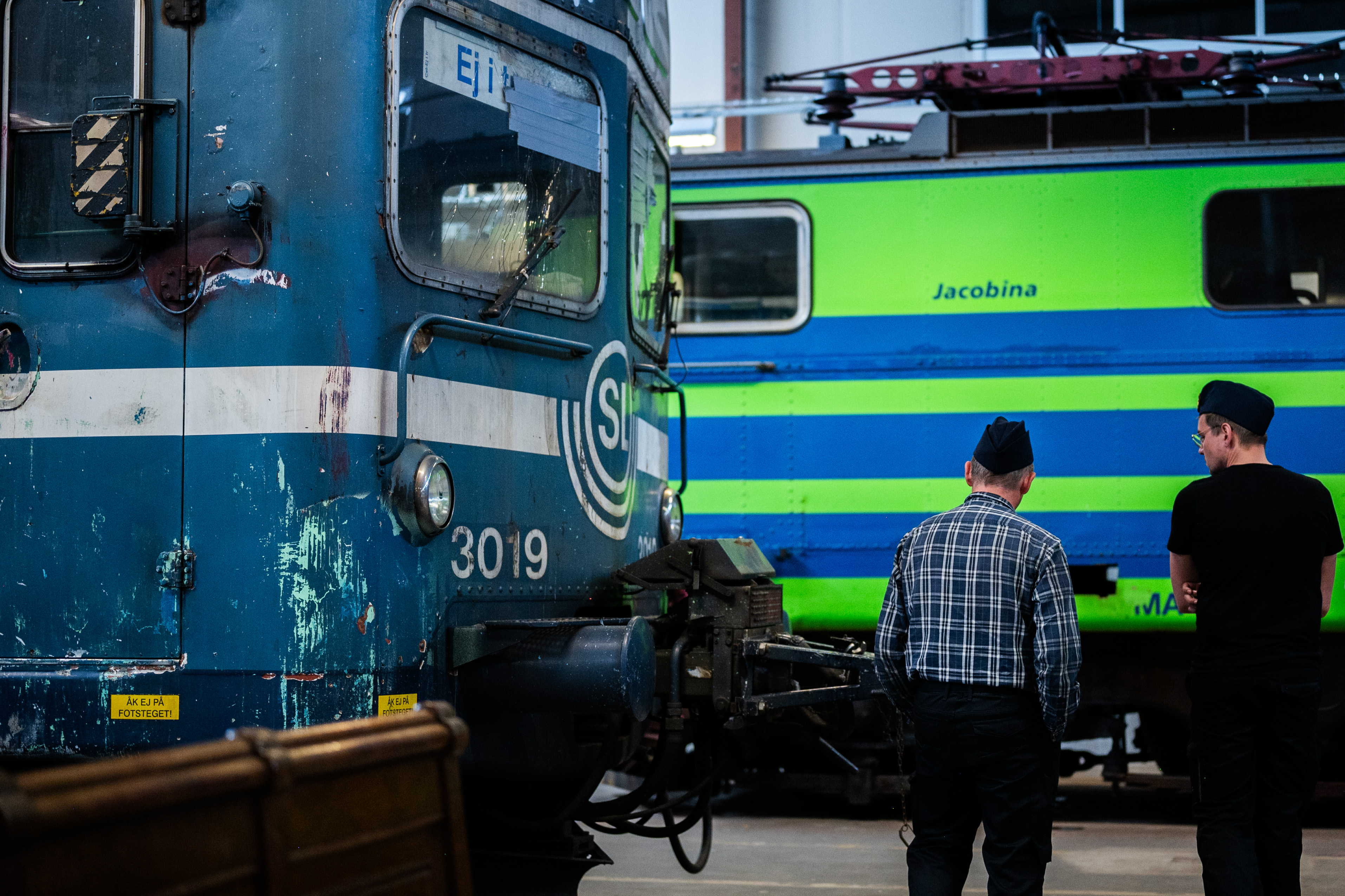 En blå tågvagn med SL loggan syns till vänster i bild. till höger står två män och kollar på en grön/blå vagn som skymtas i bakgrunden.
