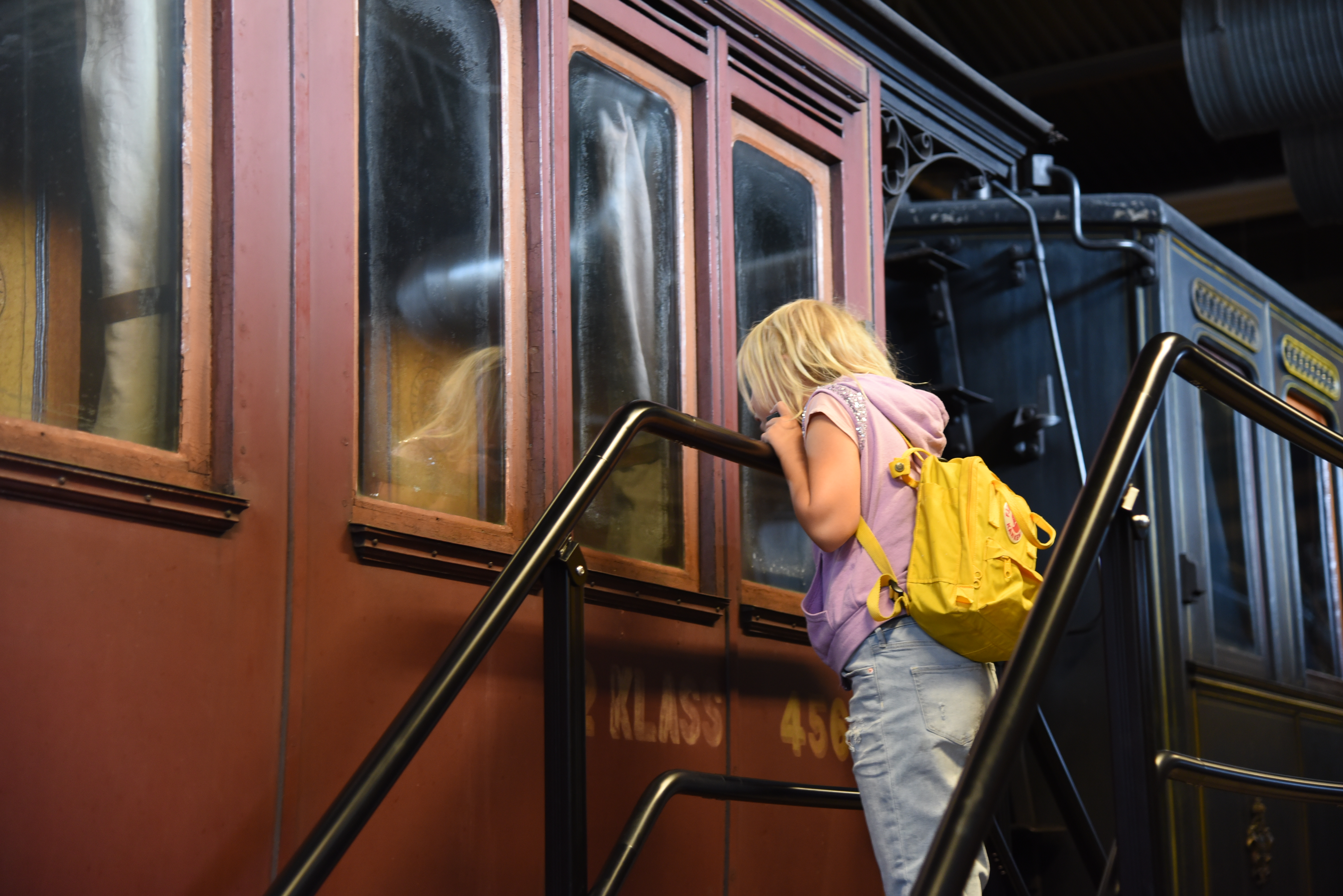 en blond flicka med rosa tröja och gul ryggsäck tittar in i fönstret på en mörkröd tågvagn.