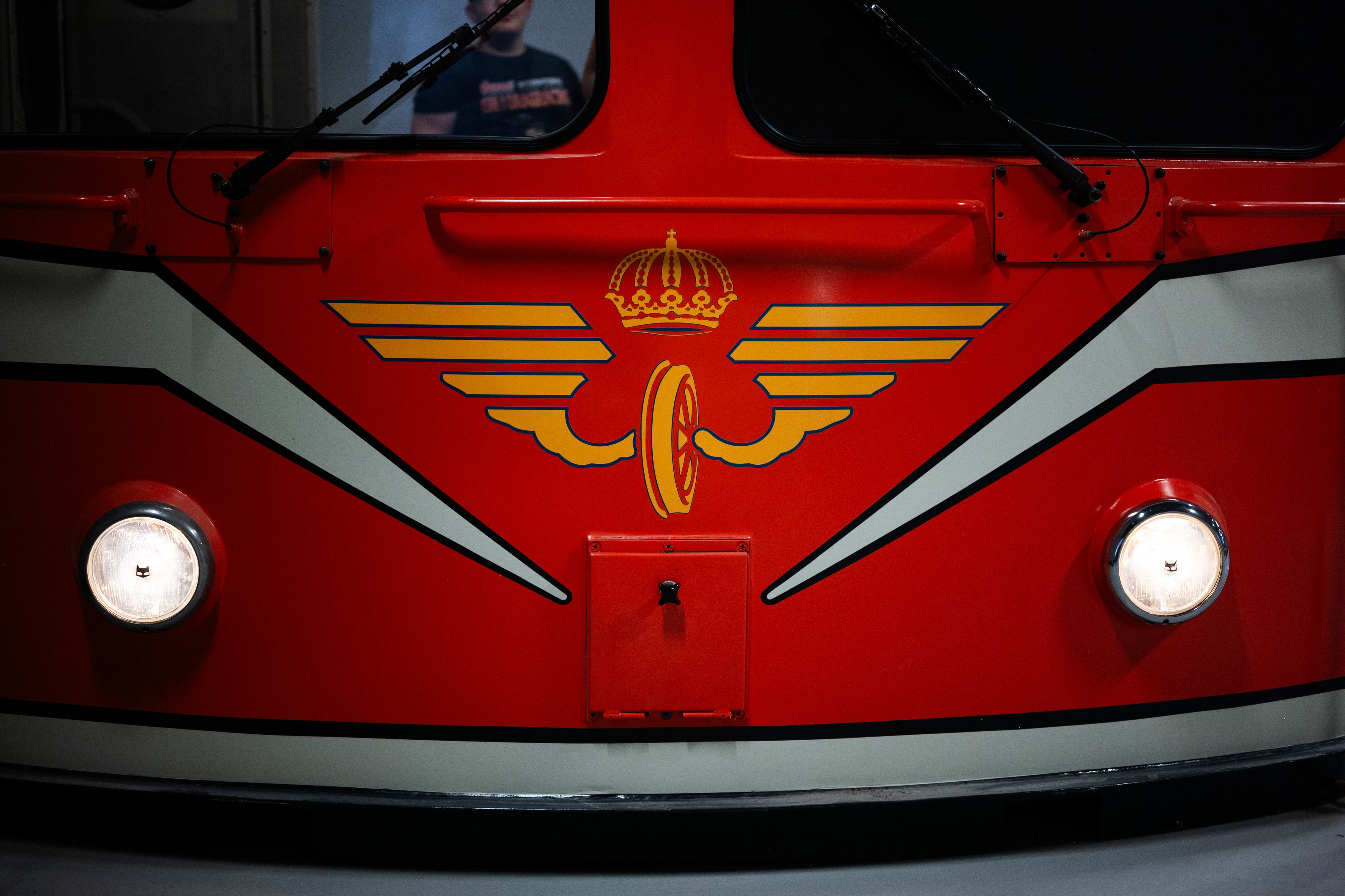Närbild på en tågvagn fram ifrån. Tågvagnen är röd med vita detaljer och två lysande lampor.