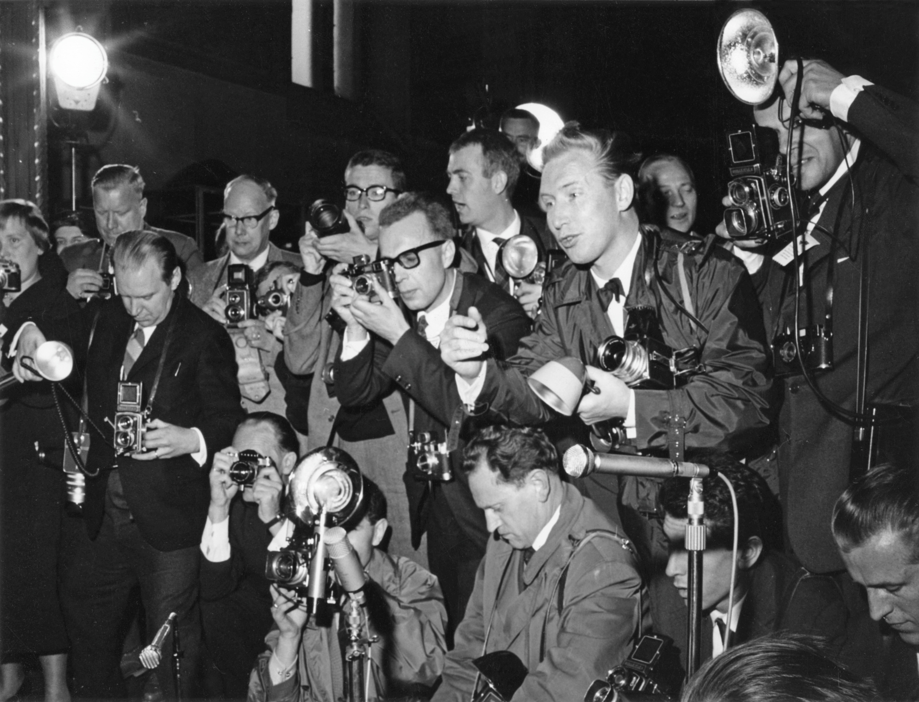 Black and white photo with a crowd of journalists dressed in suits crowding with their flashes and cameras at a press conference..