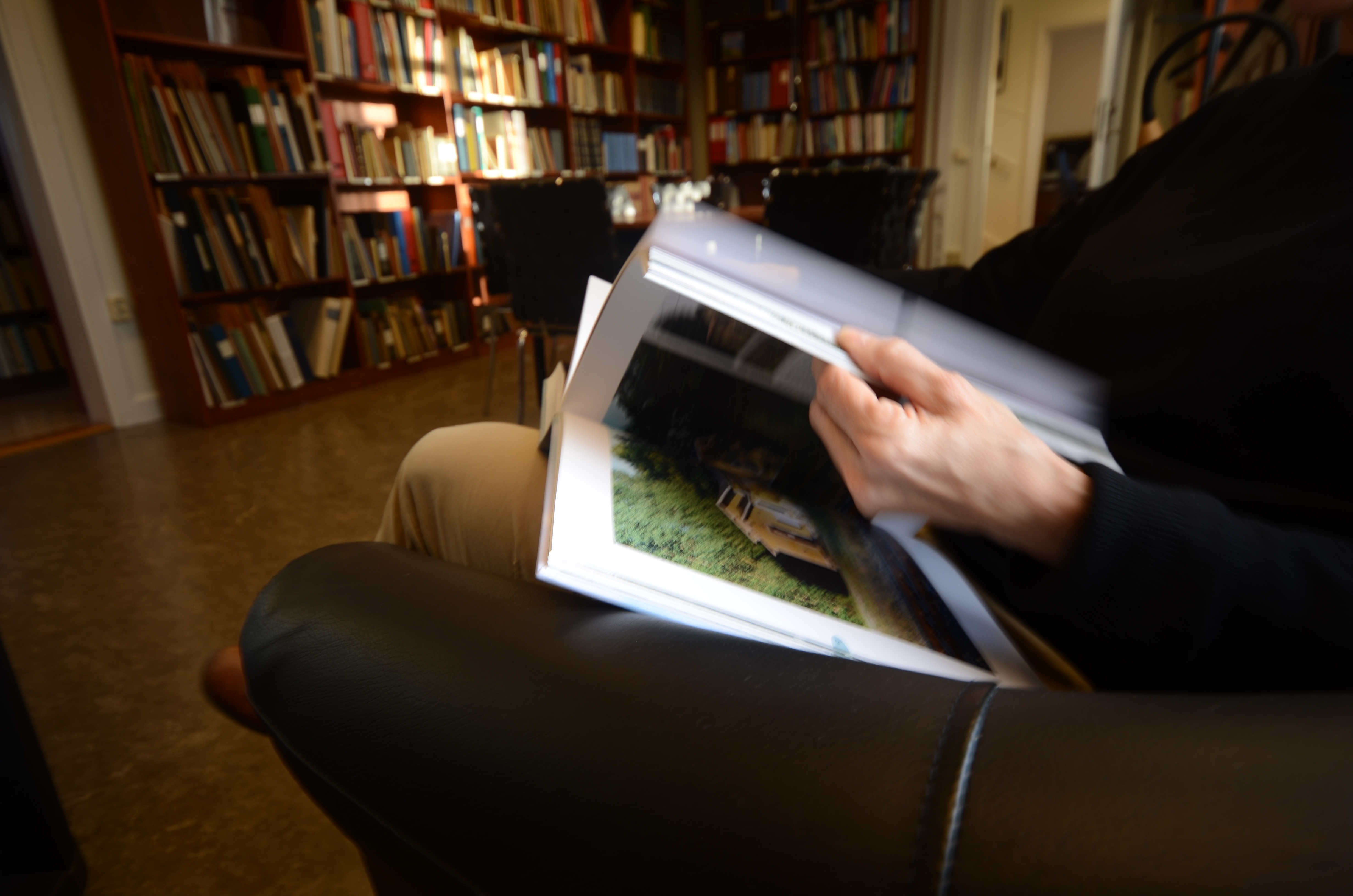 En sittande person i en skinnfåtölj bläddrar i en bok med hyllor av böcker framför sig.