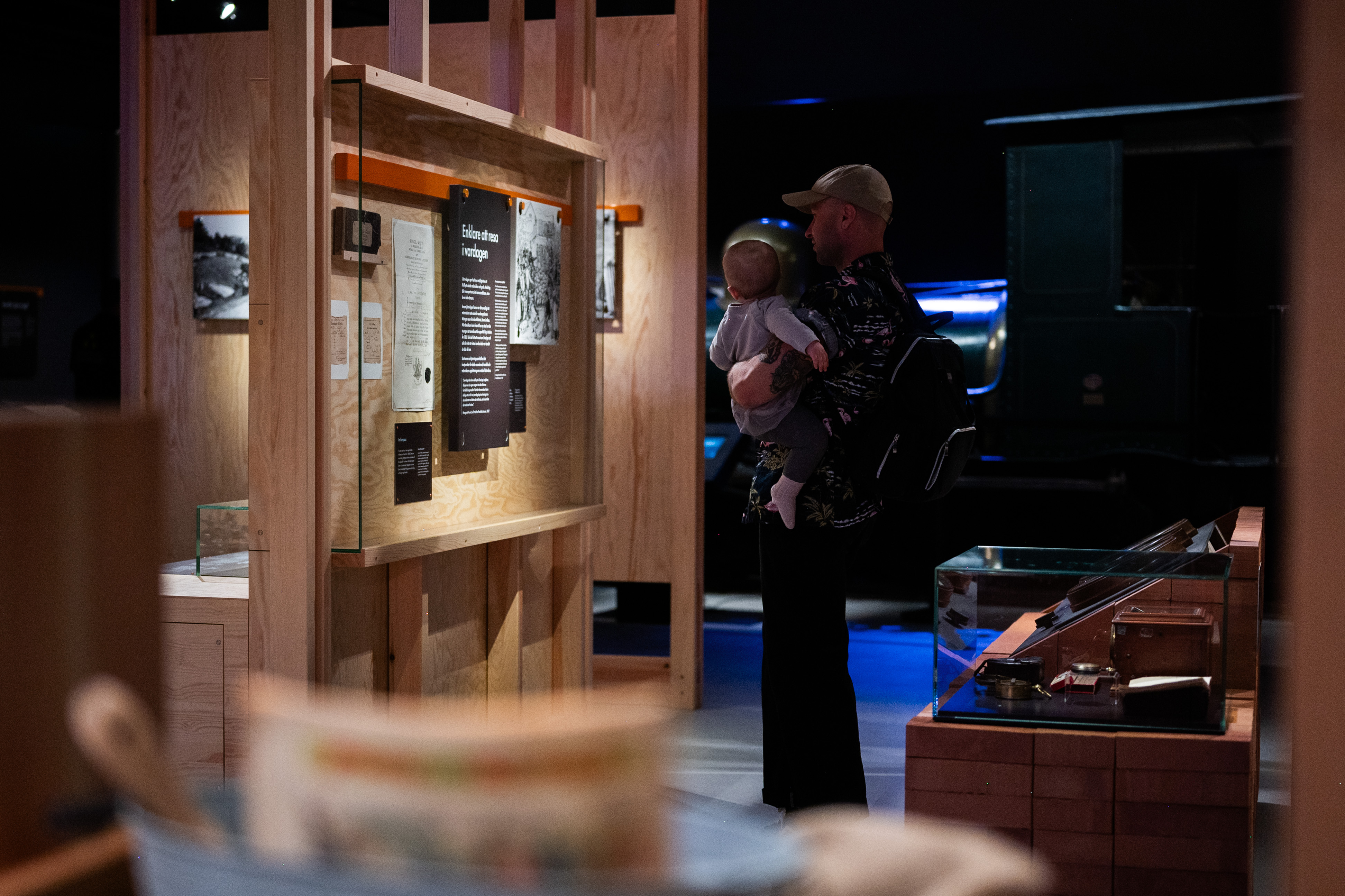 A man is holdning a baby and standing in front of a exhibition display, they are both looking at the displayed objects.