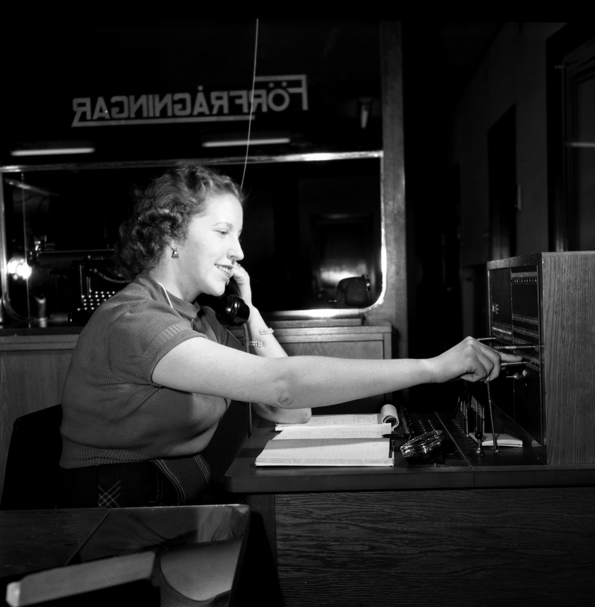 En leende växeltelefonist i kortärmad jumper och välfriserad lockad frisyr tar emot ett samtal och kopplar vidare.