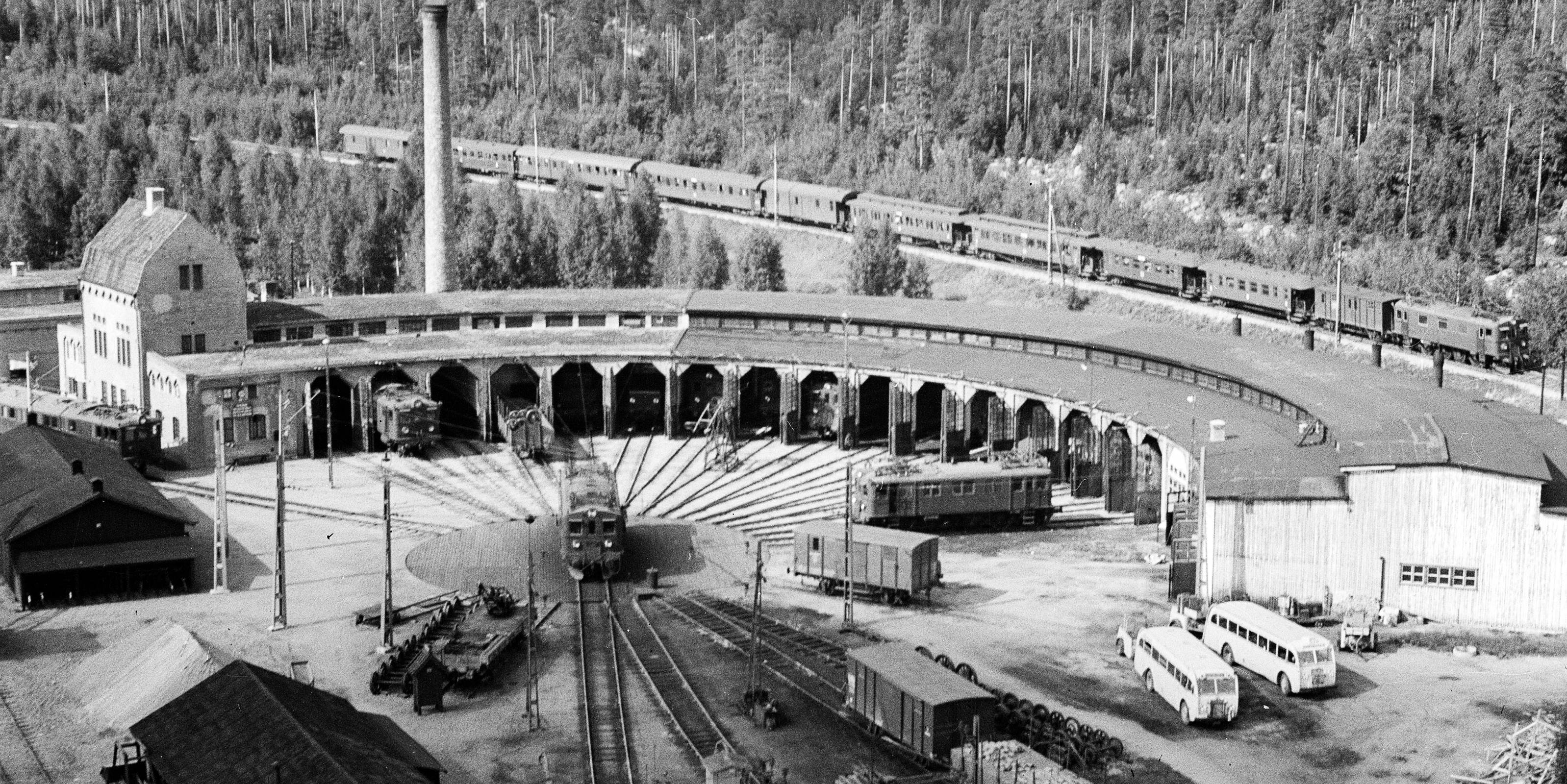 Flygfoto av större tågstall med järnväg i bakgrunden där ett tåg åker. Vagnar och lok både i stallet och utanför på räls.