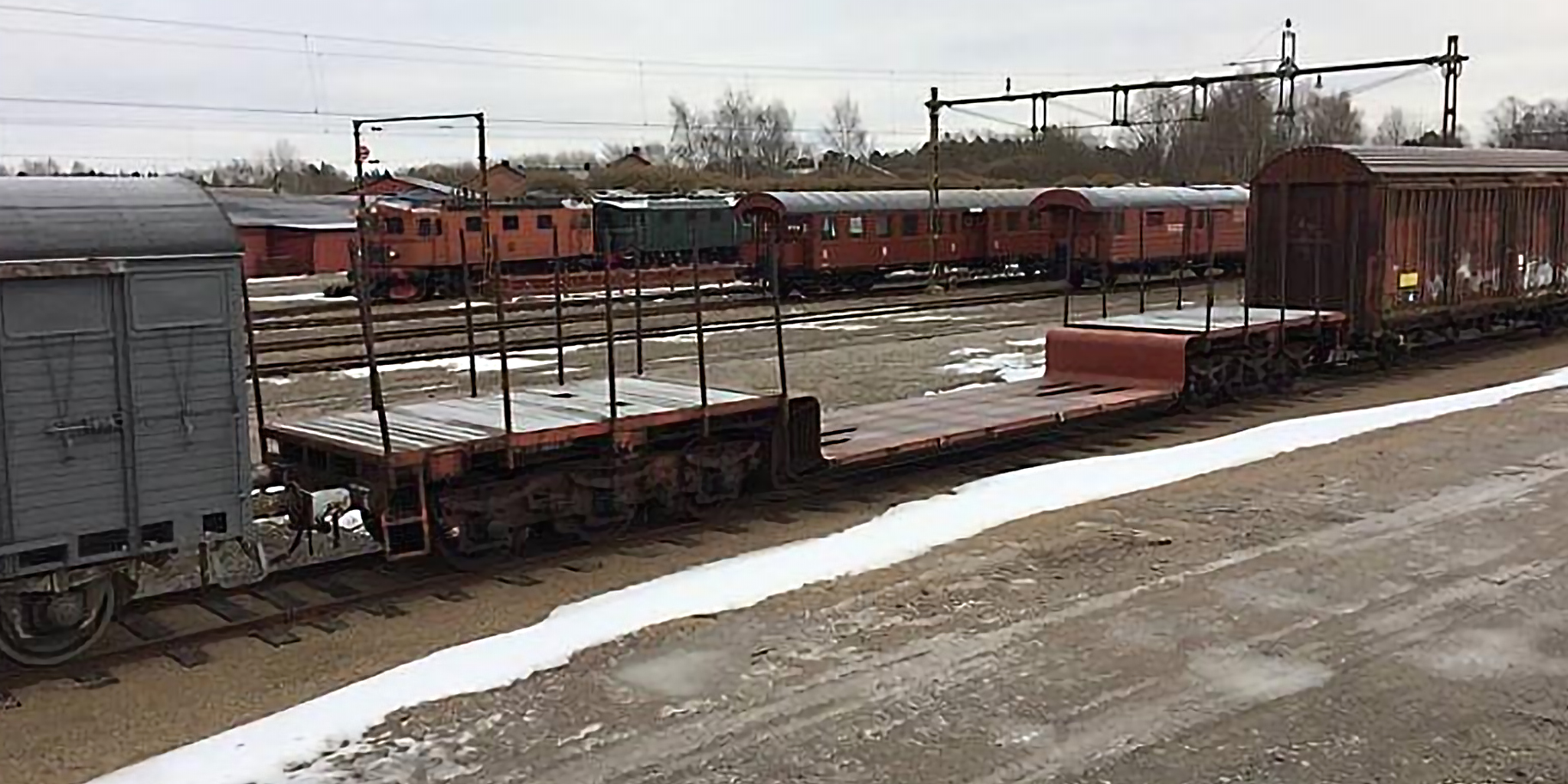 A red brown six-axle open freight car on a track area surrounded by other train cars and a streak of snow on the ground in front.