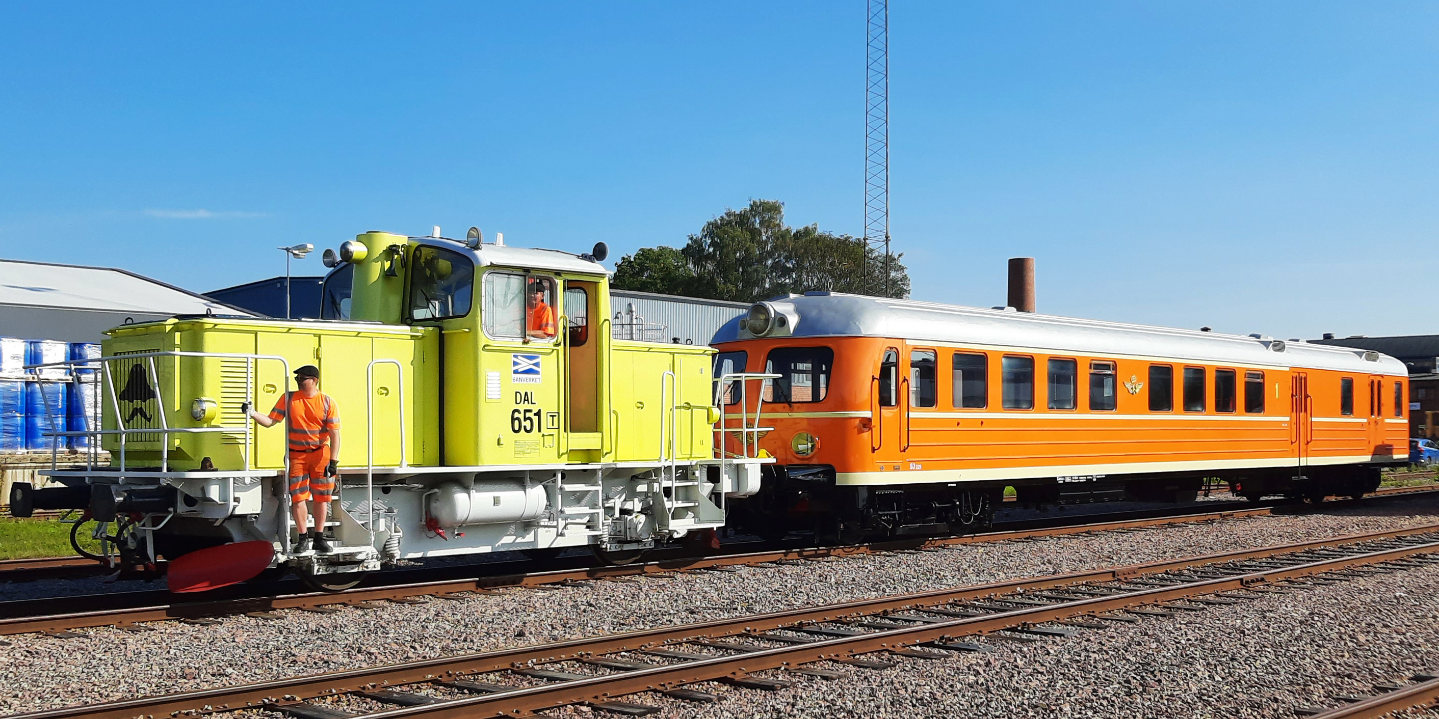 Lime yellow locomotive with orange rail bus attached. A man in the cab, a man standing at the front of the locomotive.