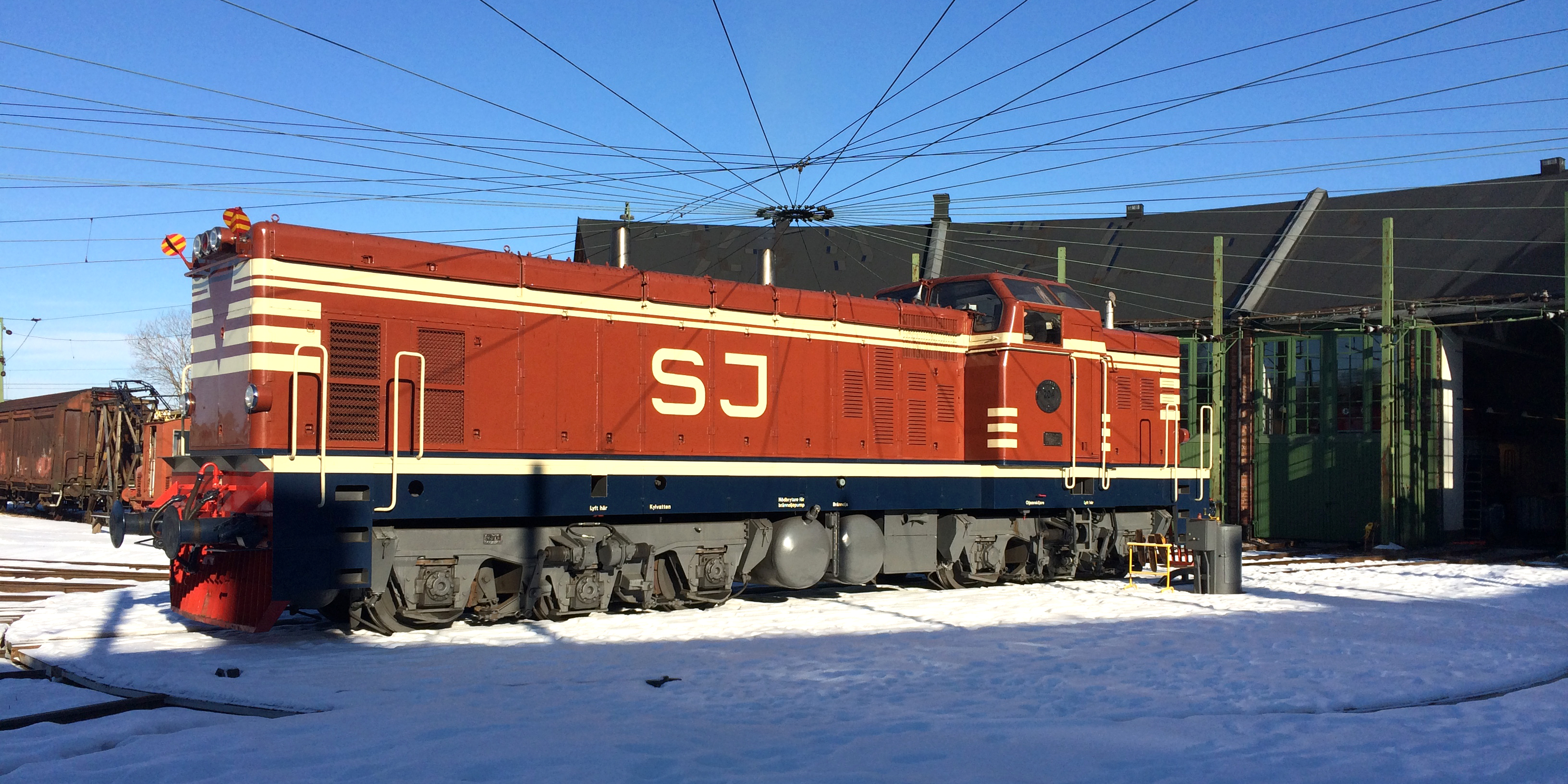 Red locomotive with the word SJ and stripes painted in white. Standing on the turntable outside a round house in a wintry landscape.