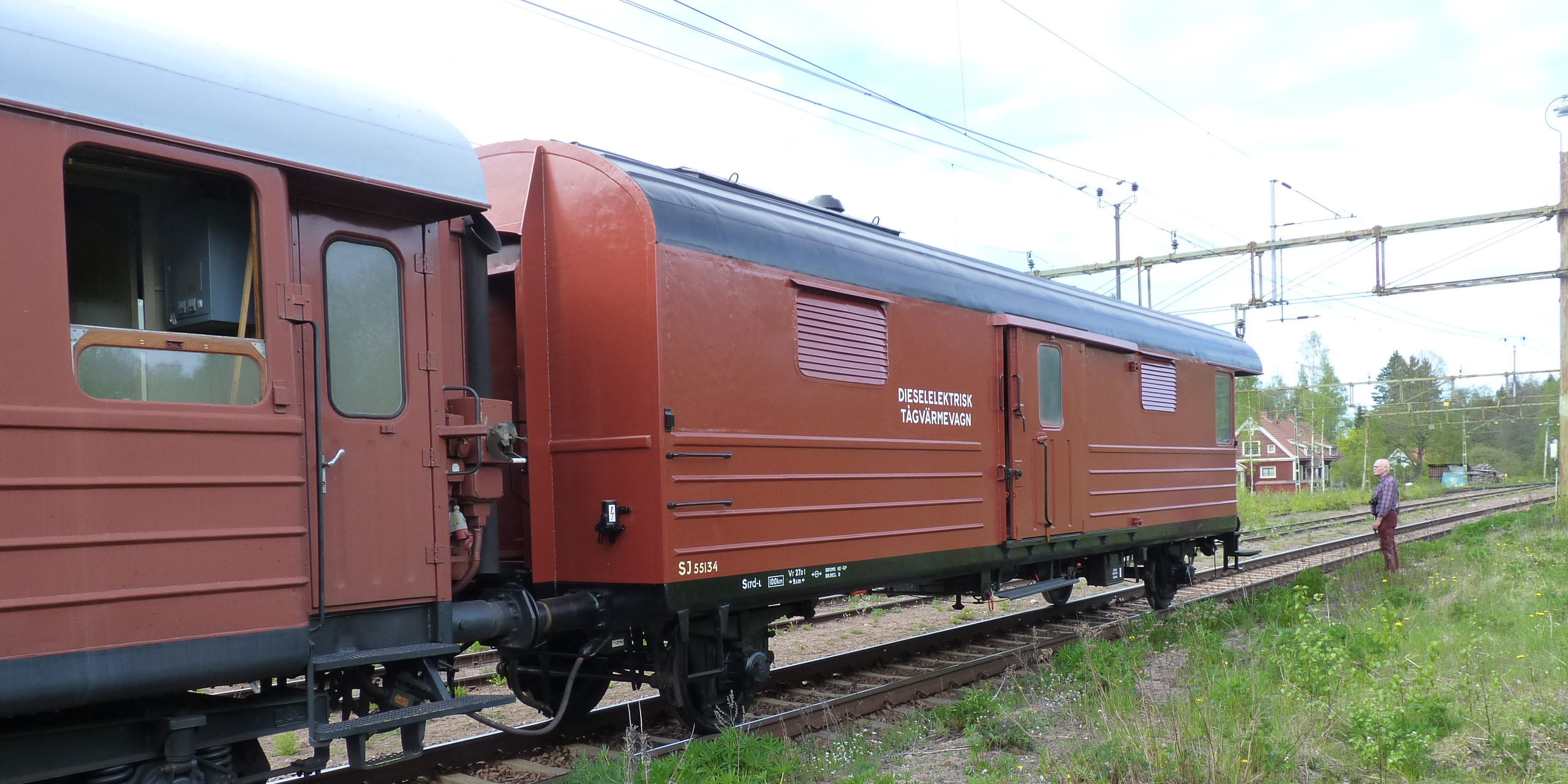 An elderly man stands by a railway track and looks at a red diesel-electric train carriage.