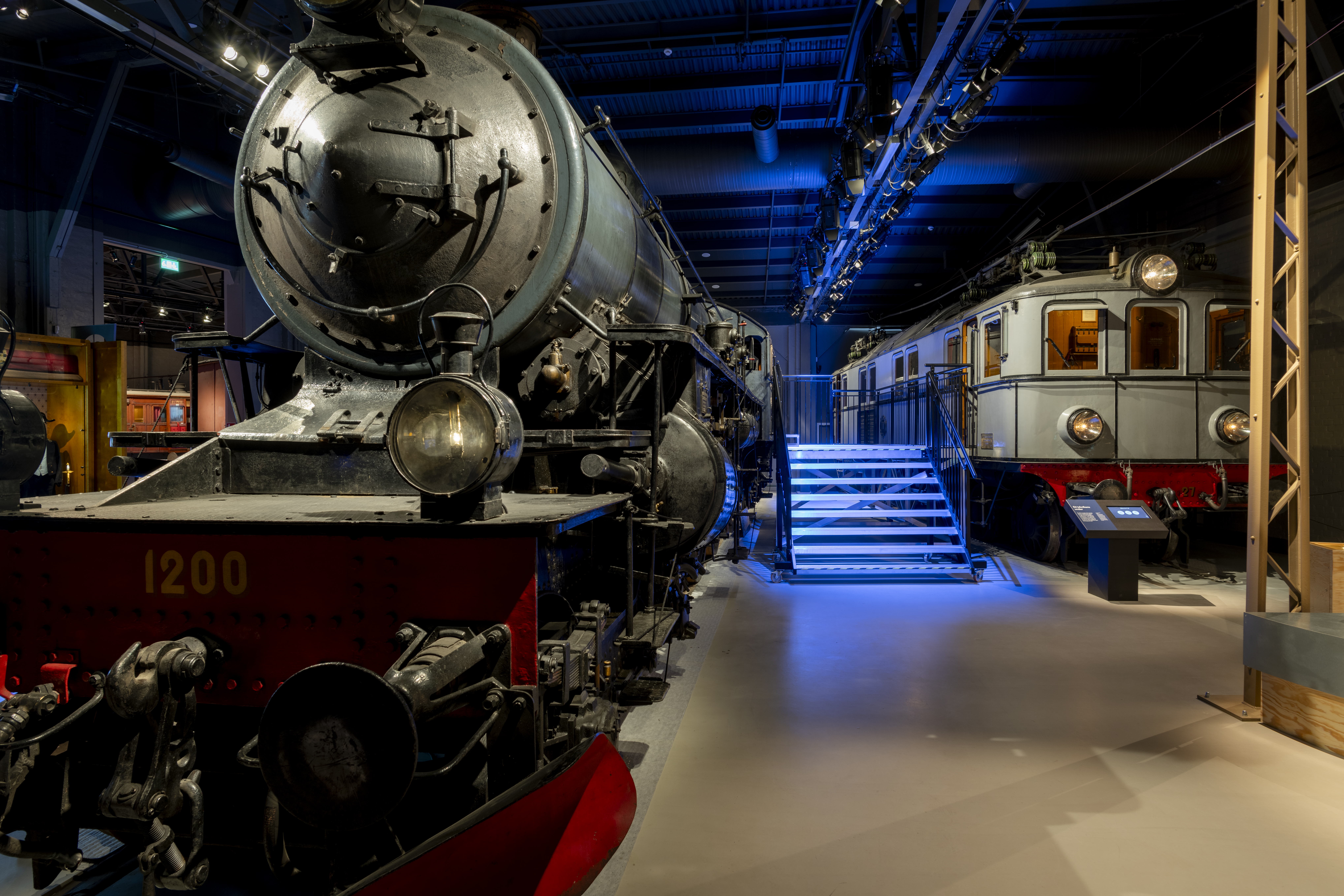 To the right there is a black steam locomotive and to the left there is a grey passanger car. In between them there are stairs so you can look into the train cars.