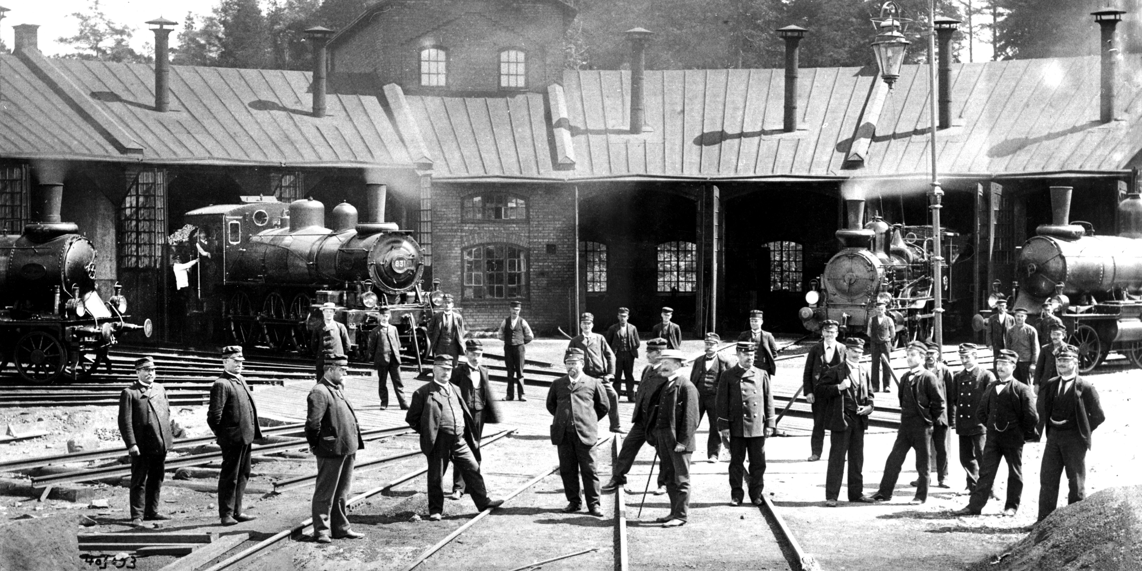 About thirty men, many in uniform, standing in front of the engine shed where several locomotives look out and puff smoke.