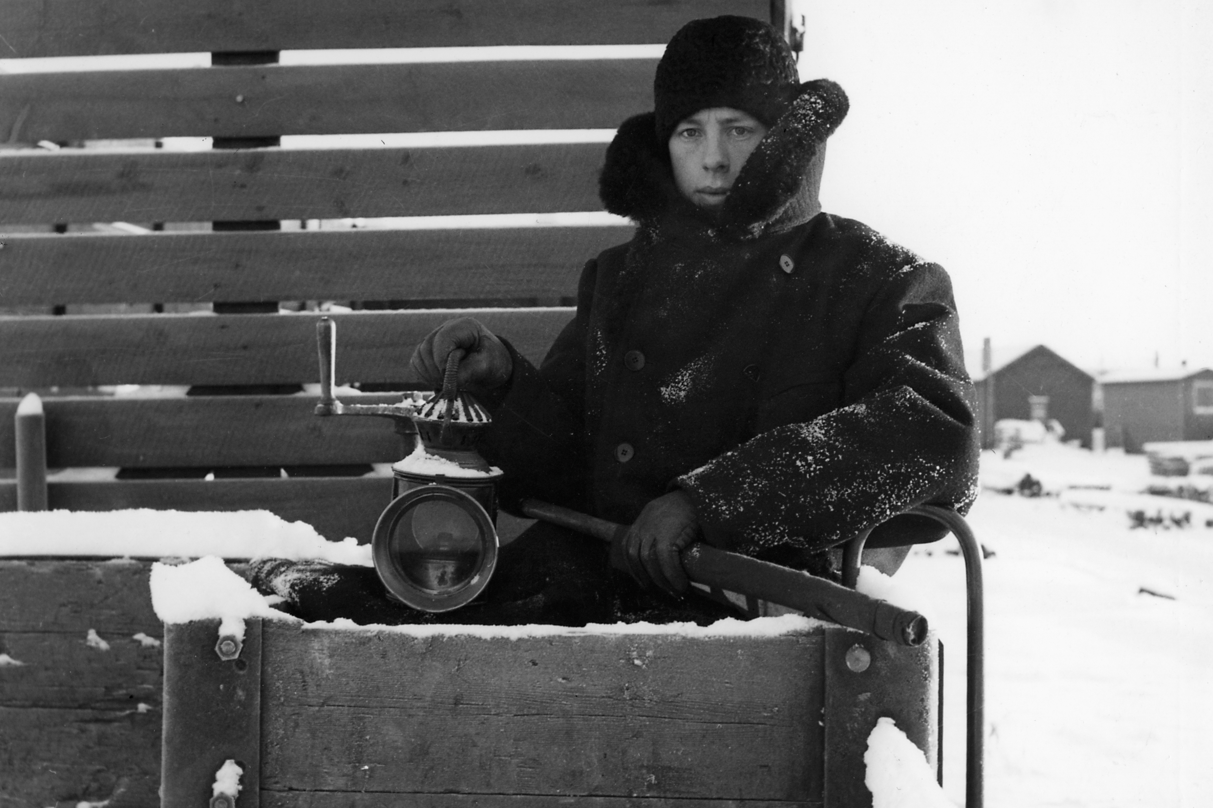 A man in a thick overcoat, hat and signal lamp in hand sits outdoors at the back of a wagon in a winter landscape.