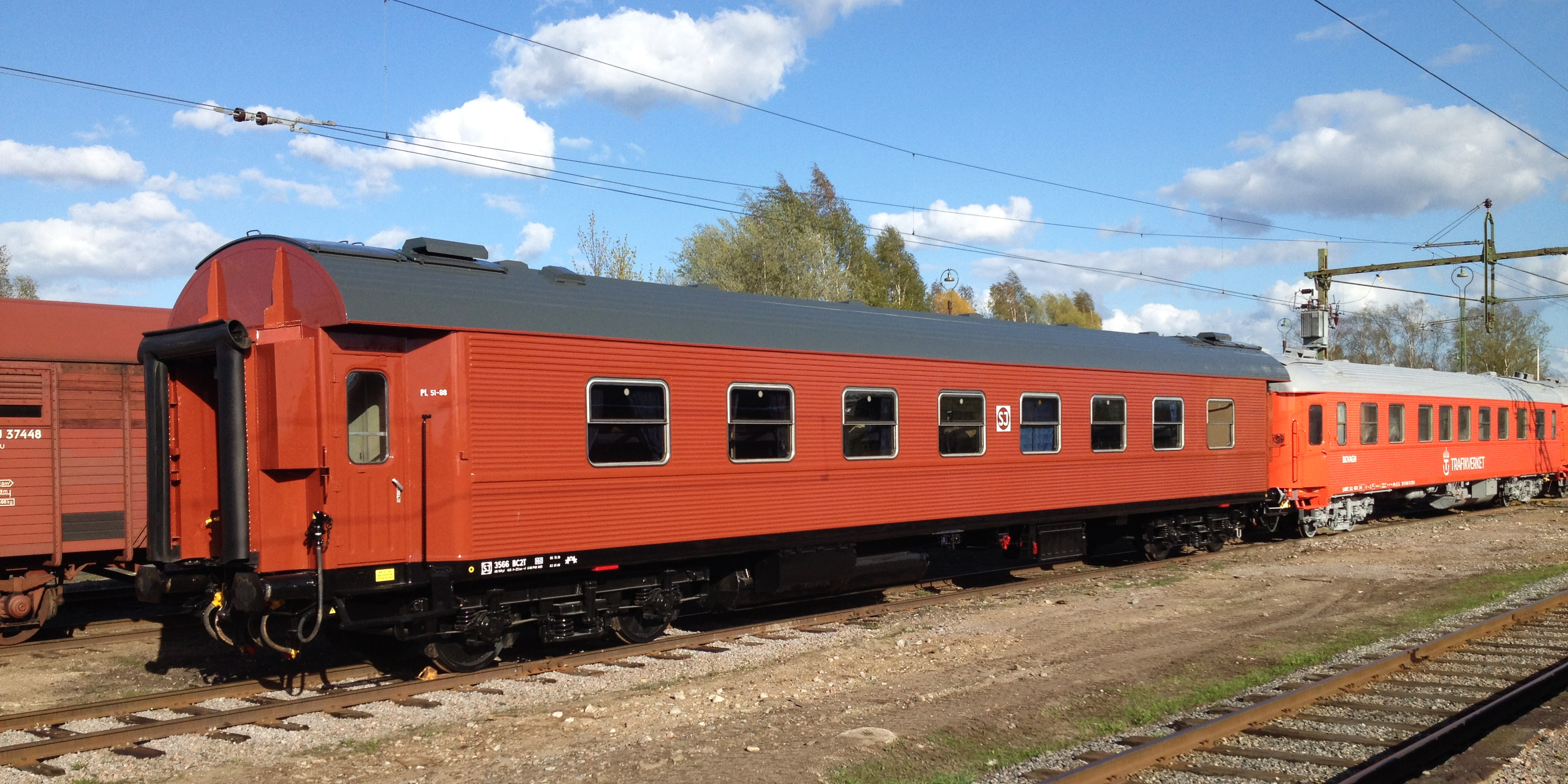 On the track, a red SJ train carriage with a lighter orange carriage following it. On the parallel track behind you can see a freight train.
