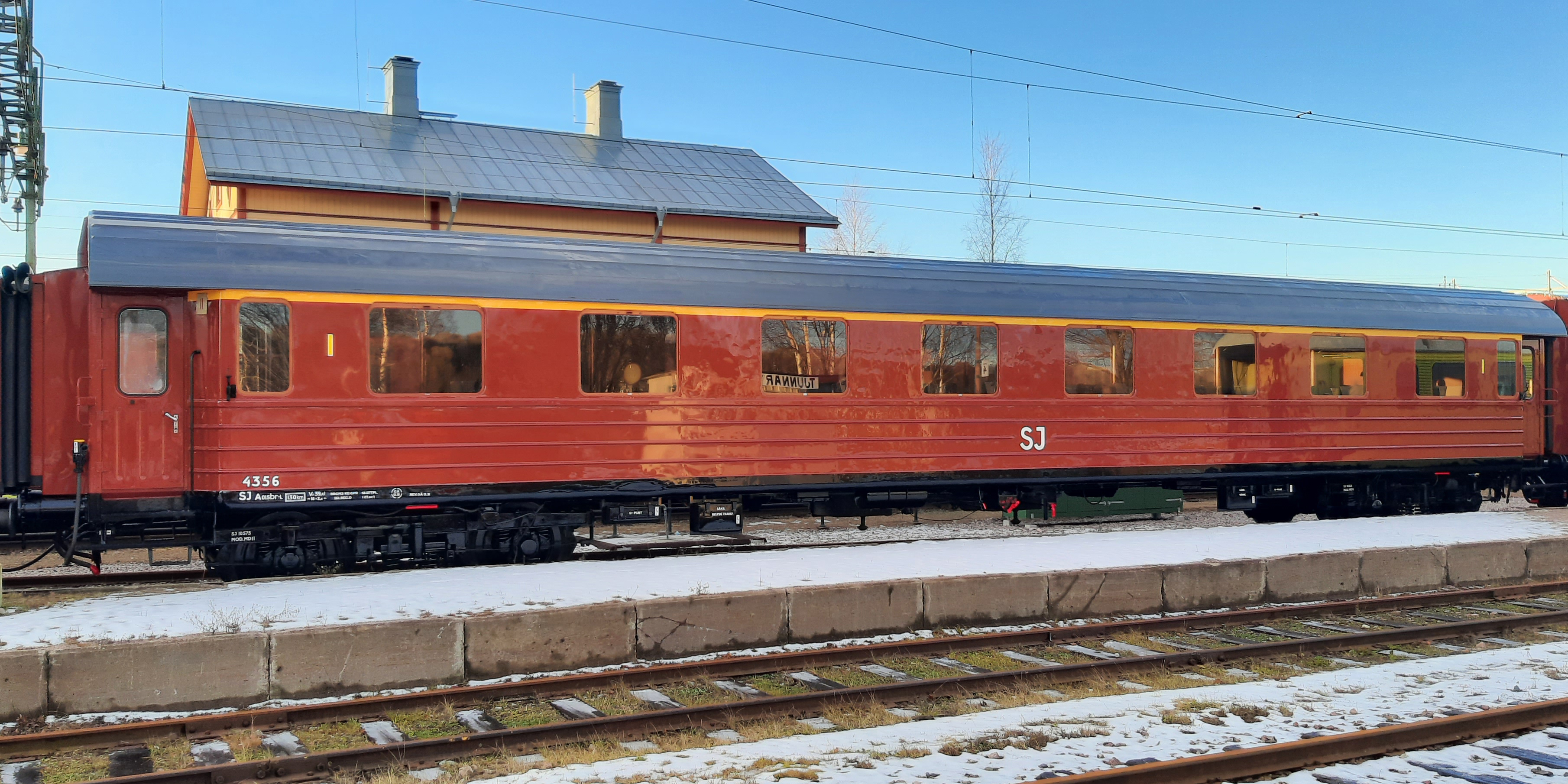 Rust red passenger car with yellow line above the windows connected between other cars on the track area.
